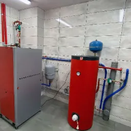 Czerwono-szary piec na paliwo stałe marki Defro z podajnikiem, zbiornik akumulacyjny ciepła w kolorze czerwonym, oraz elementy instalacji CO w kotłowni wykończonej płytkami ceramicznymi.