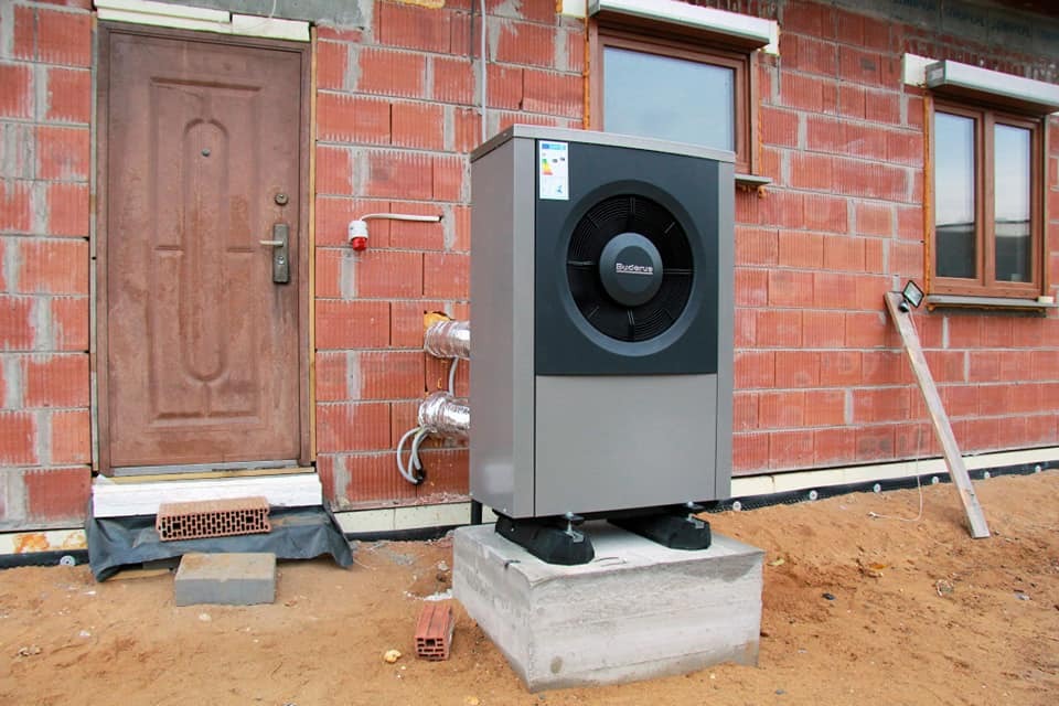 Szara pompa ciepła Buderus zamontowana na betonowym fundamencie przed budynkiem w trakcie budowy z widocznymi przewodami i ceglaną ścianą.