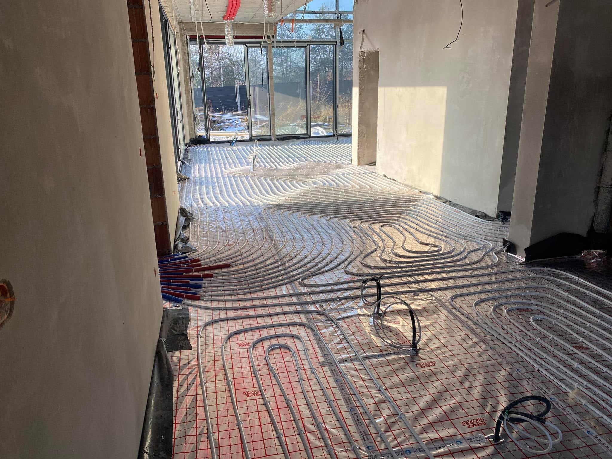 Rozłożona instalacja ogrzewania podłogowego na folii z siatką, widoczne rury w kolorze szarym i czerwono-niebieskie złączki, surowy stan wykończenia wnętrza z widokiem na zewnątrz przez duże okna.