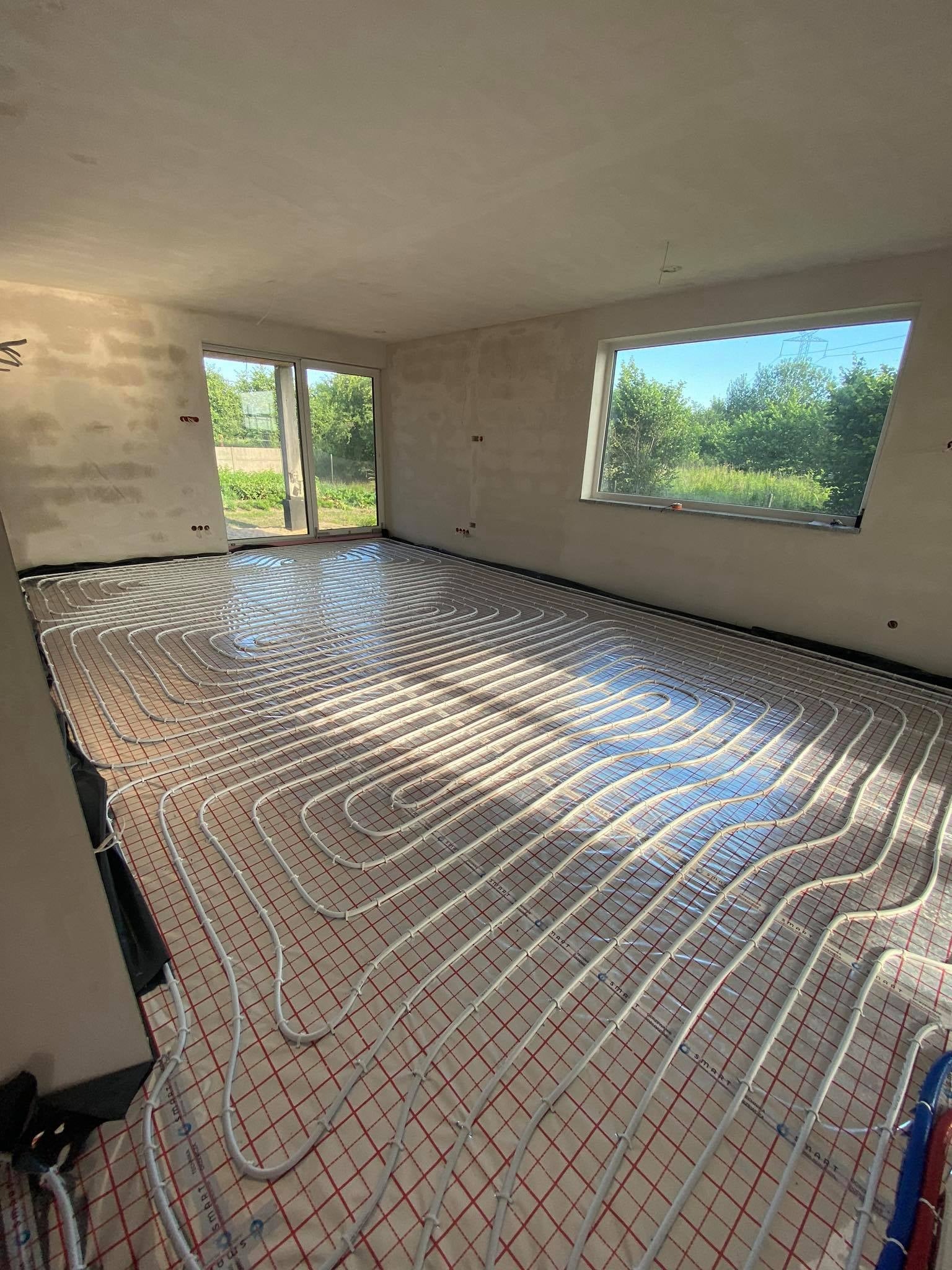Widok na podłogę z zamontowanym ogrzewaniem podłogowym w jasnym, surowym wnętrzu. Białe rury ułożone w pętle na czerwonej siatce, odbijające światło słoneczne. Okna z widokiem na zieleń na zewnątrz.
