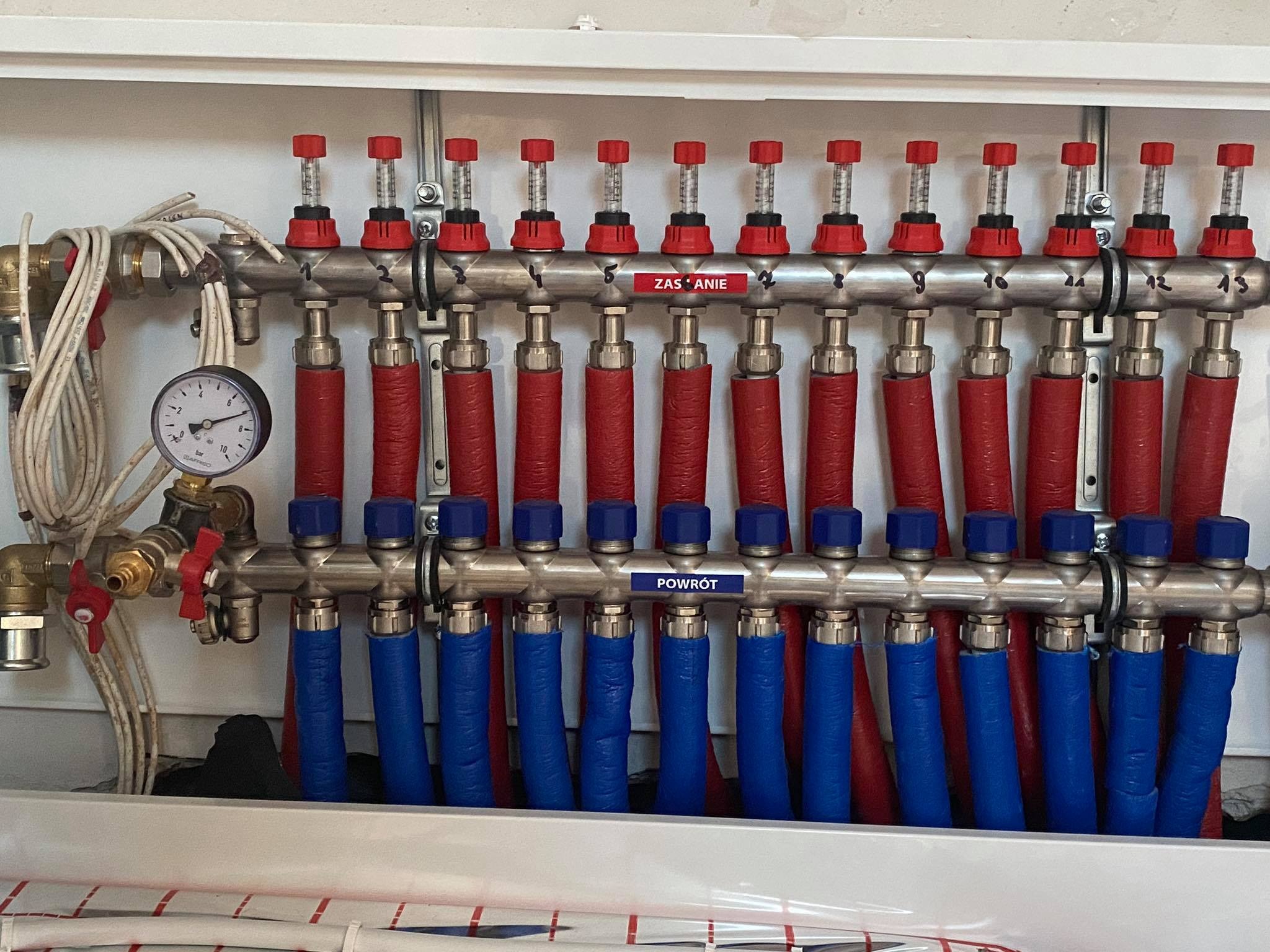 Rozdzielacz ogrzewania podłogowego z rurami oznaczonymi kolorami czerwonym i niebieskim, manometr po lewej stronie, zamontowany w białej szafce.