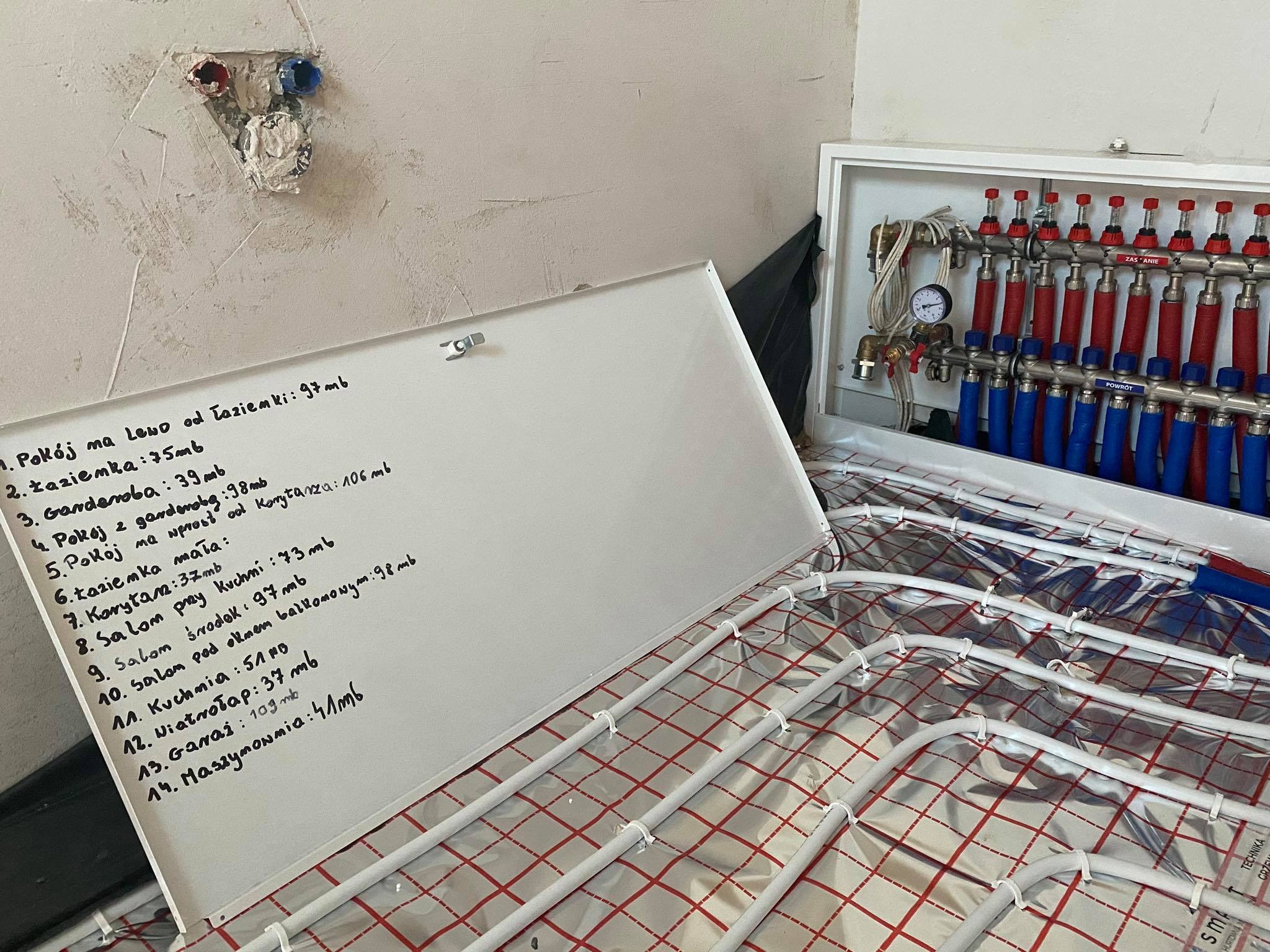 Rozdzielacz ogrzewania podłogowego z listą pomieszczeń i metrażem, widoczne białe rury na folii z czerwonymi liniami, w tle fragment ściany z instalacją hydrauliczną.