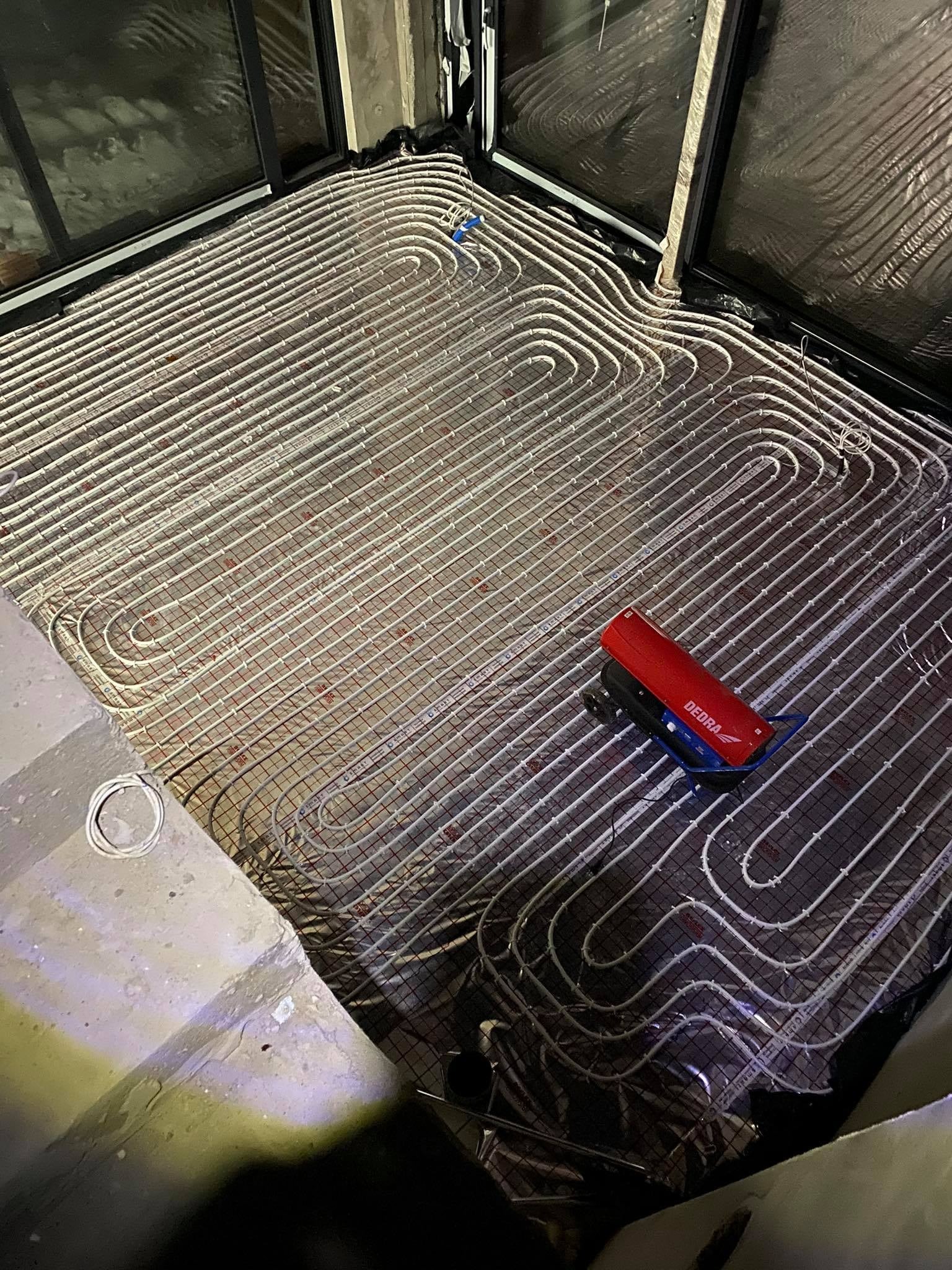 Rozłożona instalacja ogrzewania podłogowego z rurkami na siatce, widoczny fragment ściany i okna oraz narzędzie do montażu rur DEBRA.