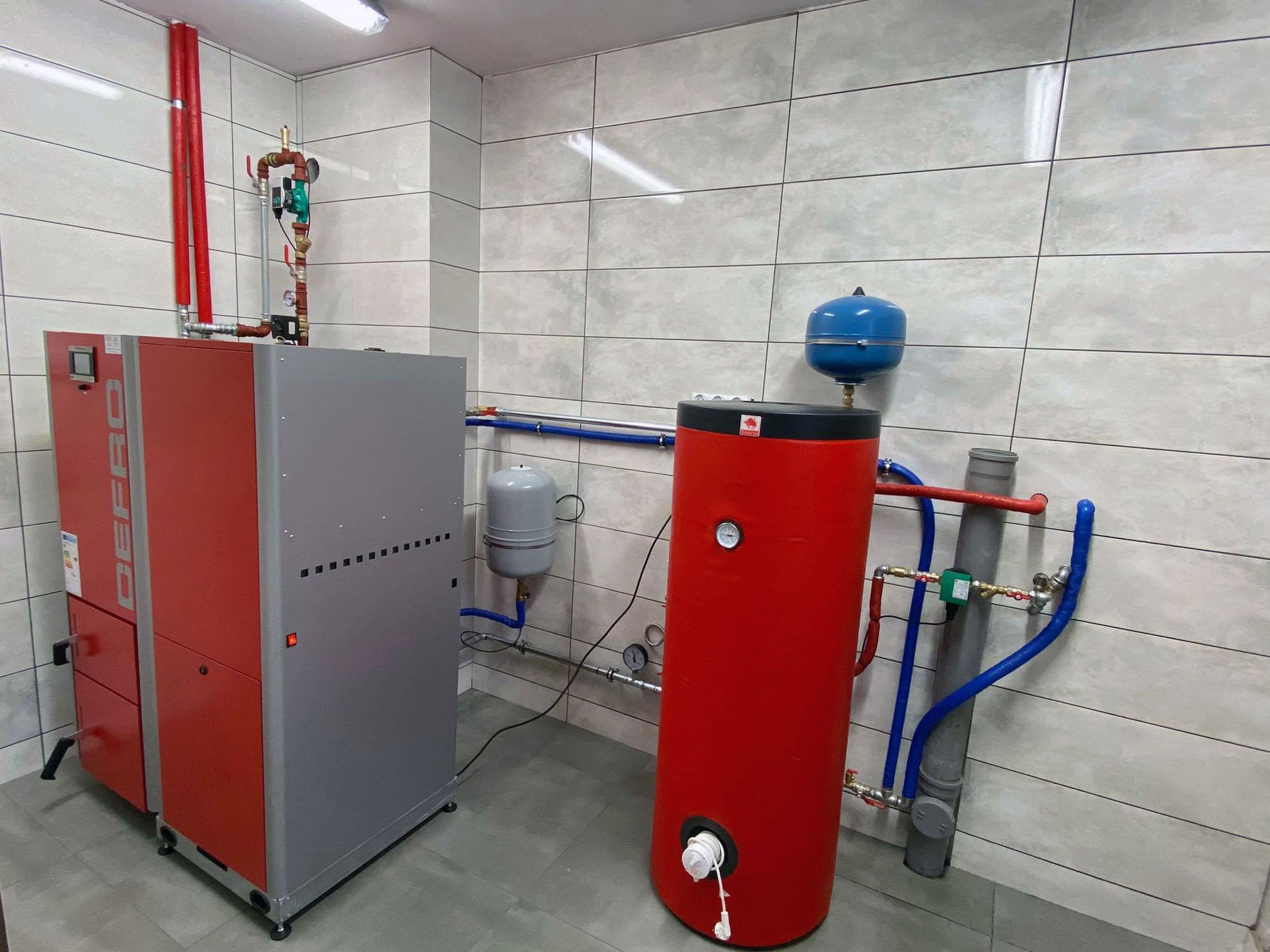 Czerwono-szary piec na paliwo stałe marki Defro z podajnikiem, zbiornik akumulacyjny ciepła w kolorze czerwonym, oraz elementy instalacji CO w kotłowni wykończonej płytkami ceramicznymi.