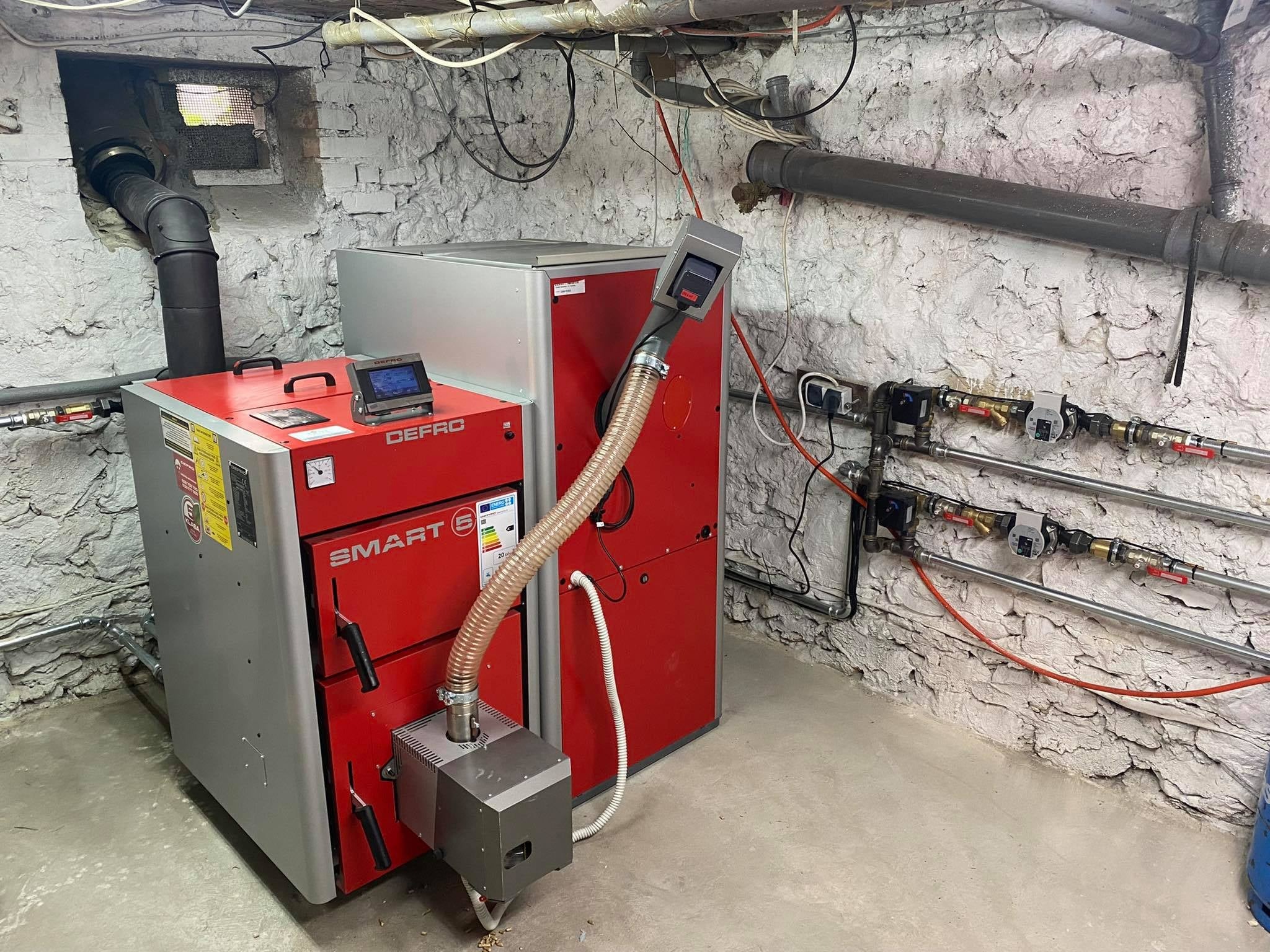 Nowoczesny piec na paliwo stałe Defro Smart S z podajnikiem, zainstalowany w piwnicy o ścianach z surowego kamienia, widoczne rury i instalacje.