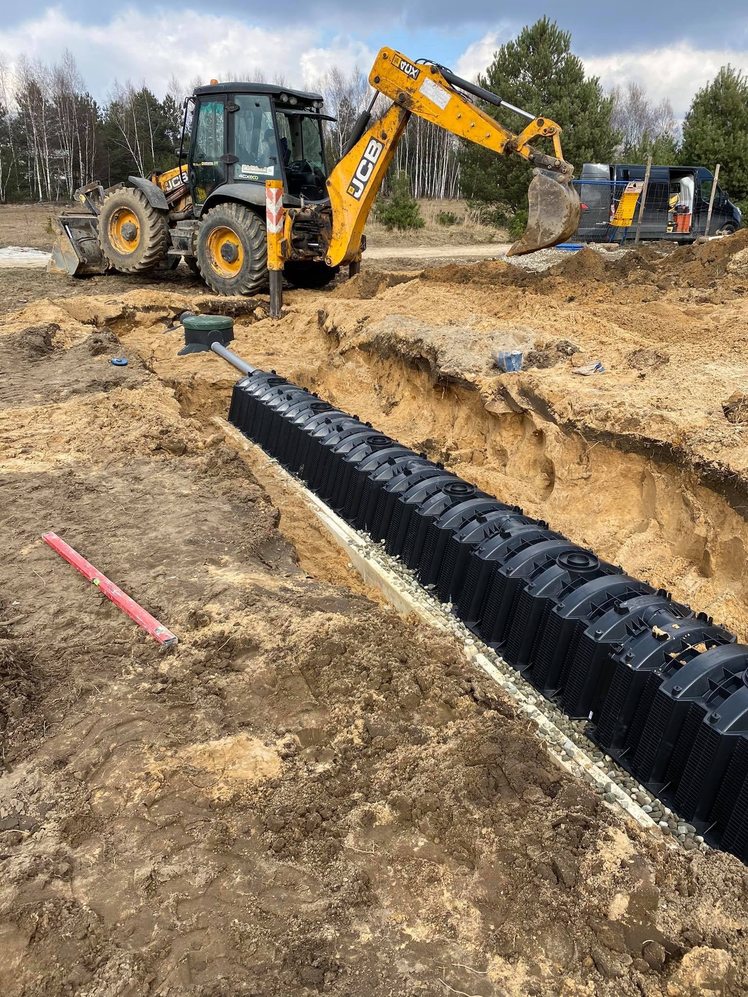 Instalacja drenażu rozsączającego z rur drenarskich w wykopie, z widoczną koparką JCB i poziomnicą w tle.