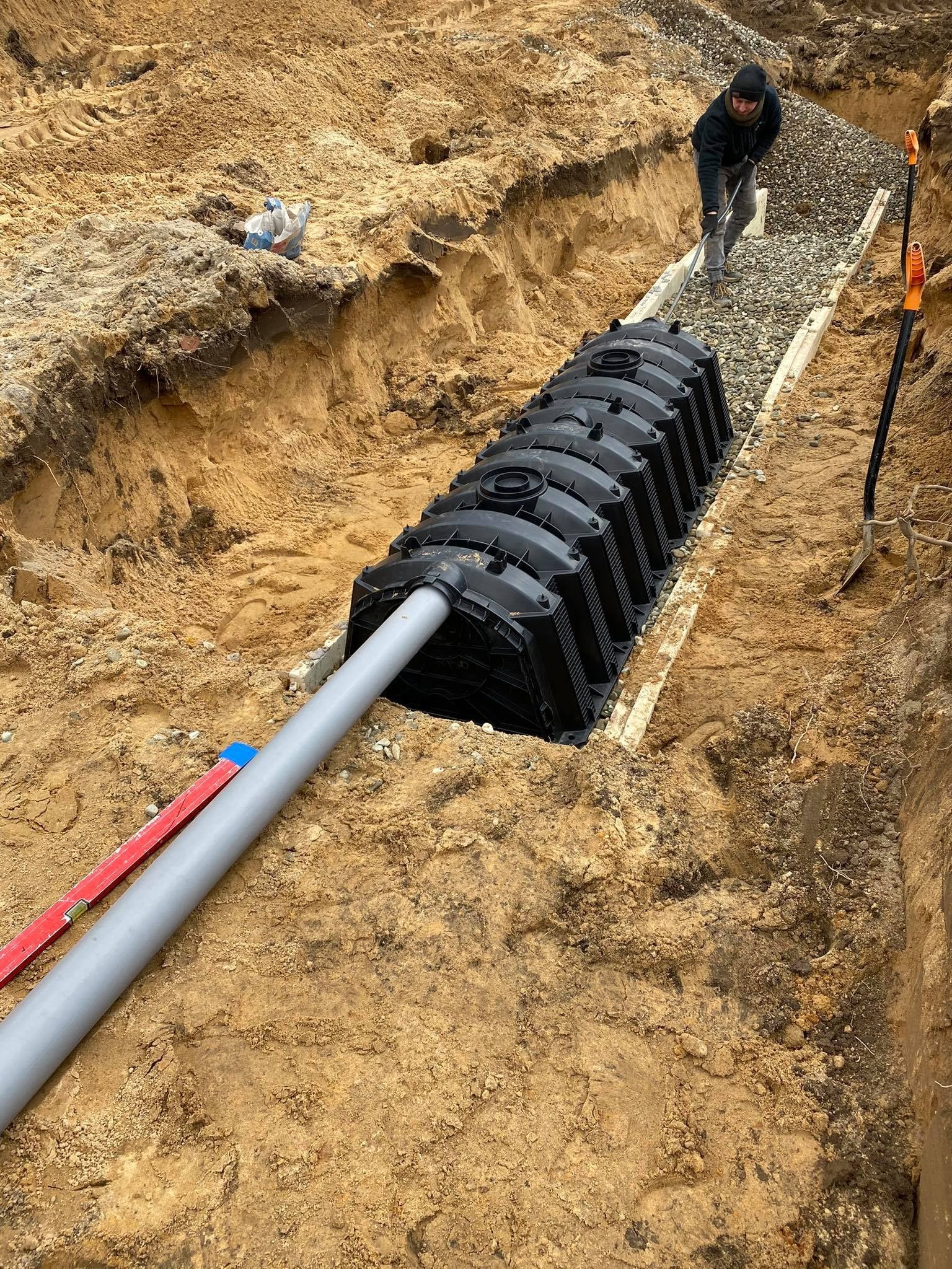 Montaż podziemnego zbiornika retencyjnego z rurą odpływową w wykopie, pracownik wyrównuje żwir, widoczne narzędzia i poziomica.