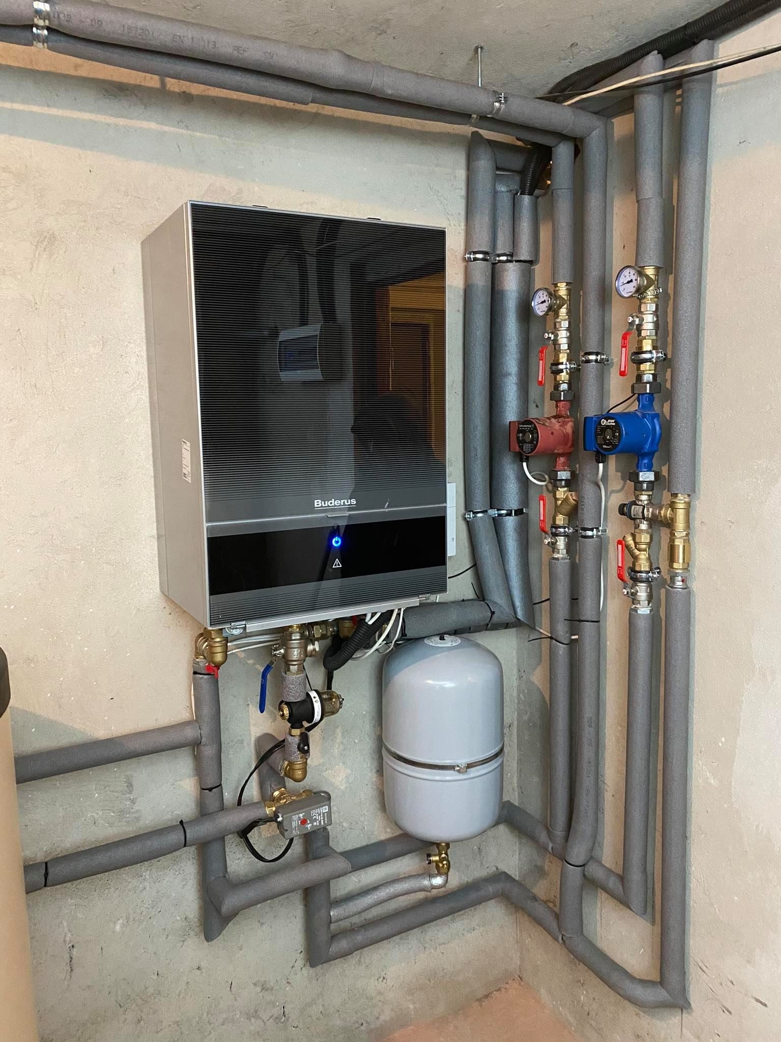 Nowoczesny, srebrny kocioł gazowy Buderus z podłączonymi rurami w izolacji, manometry i pompy obiegowe na ścianie w surowym wykończeniu.