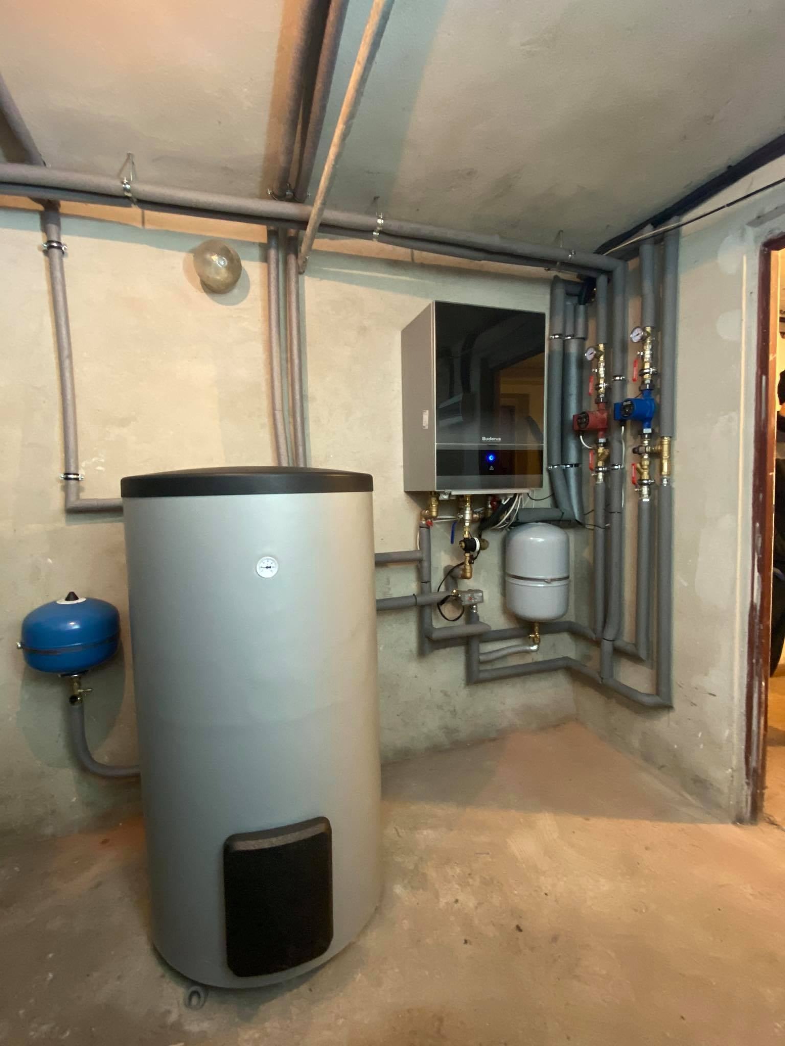 Szary zbiornik na wodę obok zamontowanego, nowoczesnego pieca gazowego Buderus z licznymi rurami i zaworami w piwnicy z surowym betonowym sufitem.