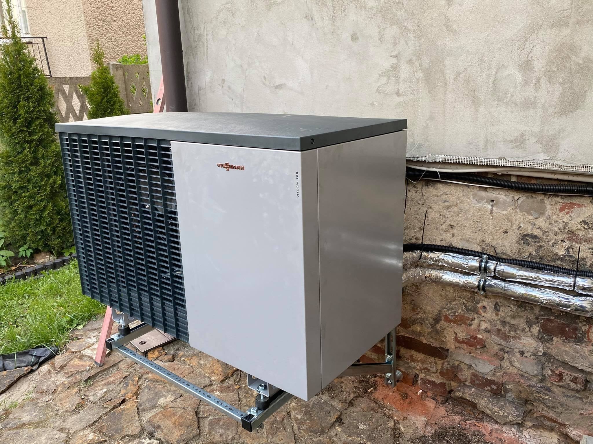 Zewnętrzna jednostka pompy ciepła Viessmann Vitocal 200 na metalowym stelażu, zamontowana przy ścianie z widocznymi rurami w izolacji aluminiowej.