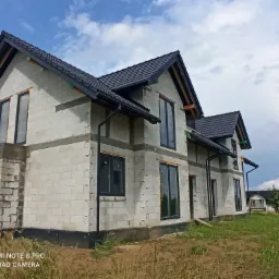 Surowy, dwukondygnacyjny dom z białych bloczk&oacute;w, z zamontowanymi czarnymi oknami i dachem z czarnej dach&oacute;wki, w trakcie budowy, na tle błękitnego nieba z chmurami.