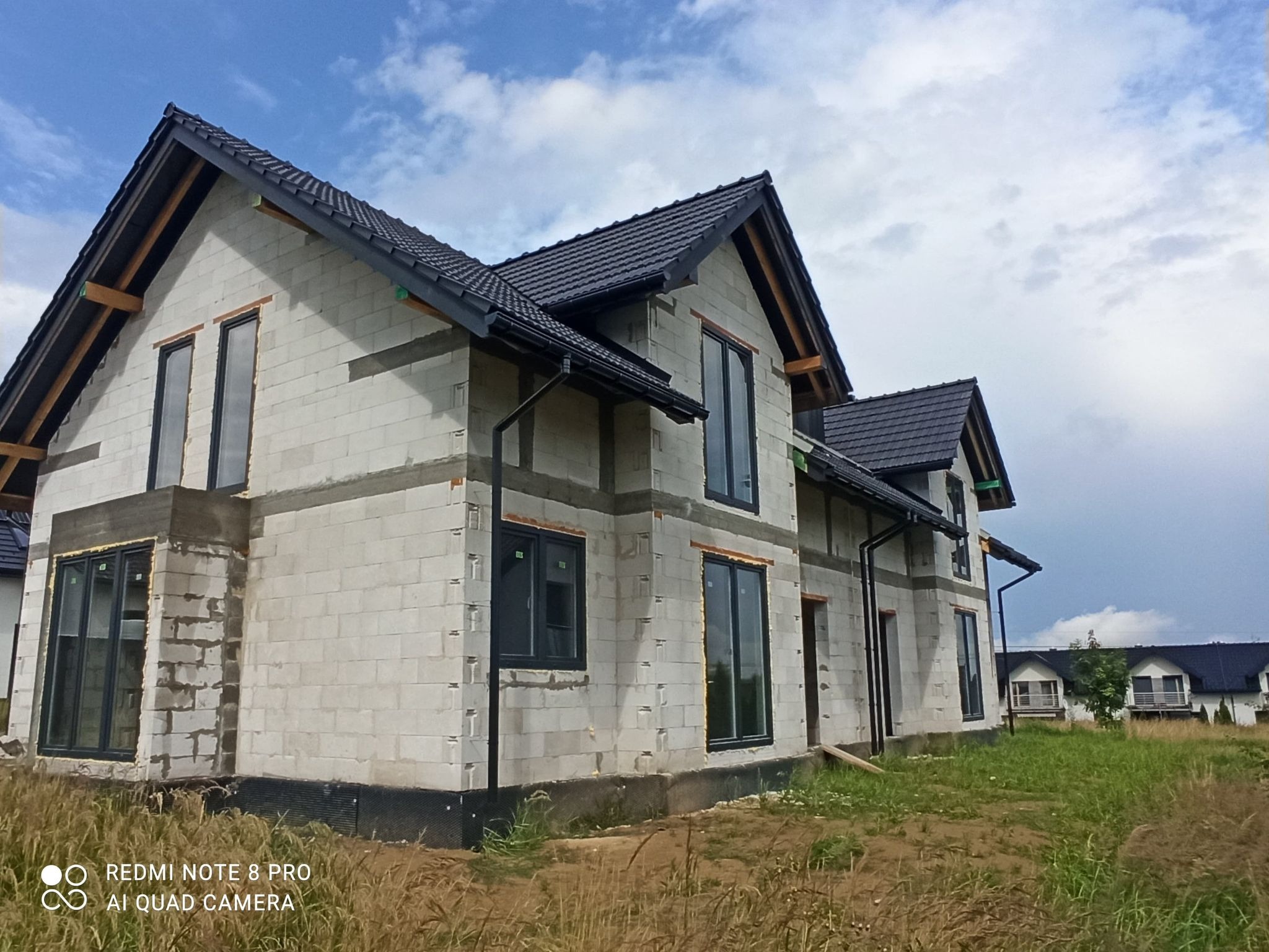 Surowy, dwukondygnacyjny dom z białych bloczków, z zamontowanymi czarnymi oknami i dachem z czarnej dachówki, w trakcie budowy, na tle błękitnego nieba z chmurami.