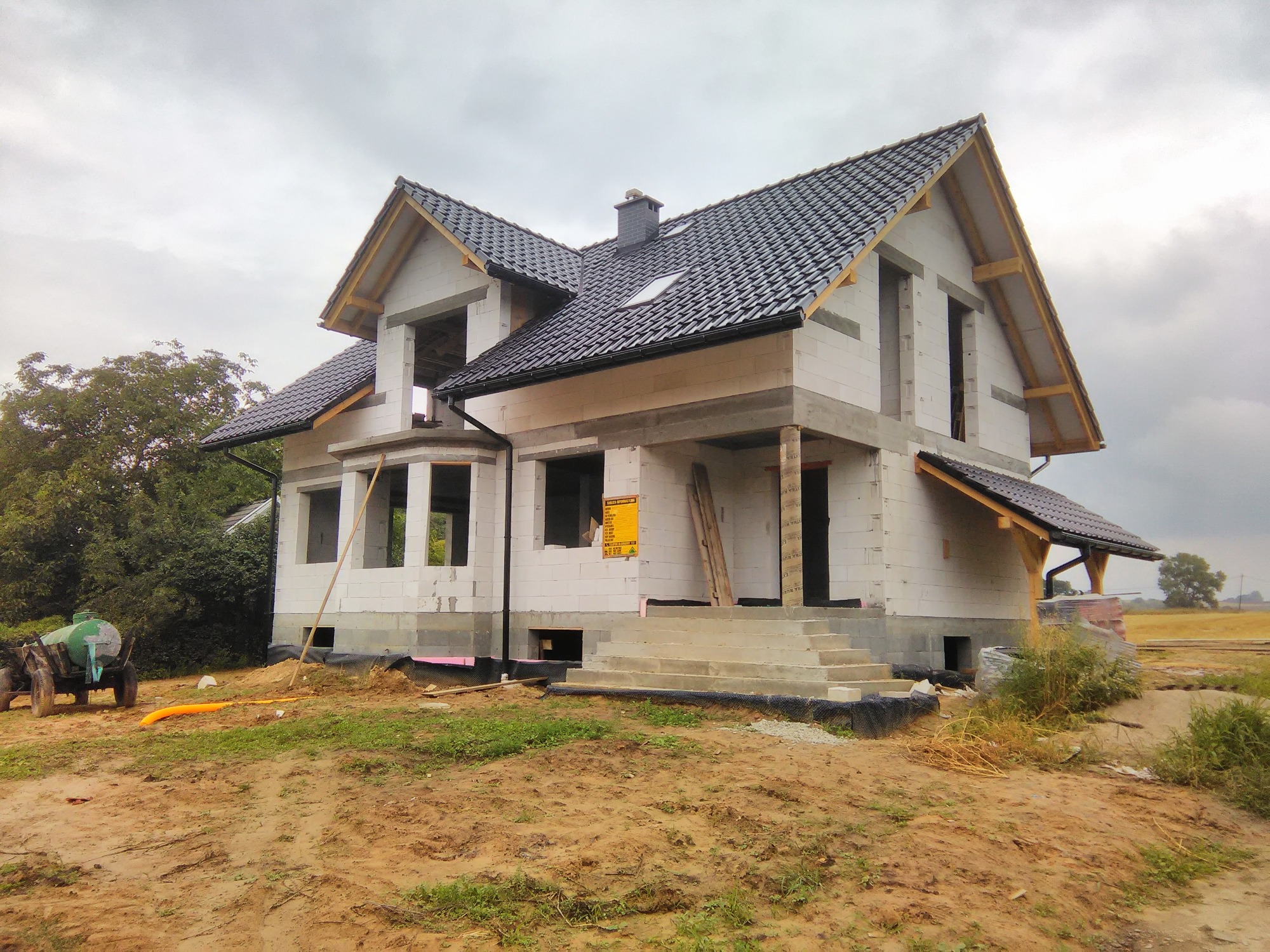 Surowy stan dwukondygnacyjnego domu z białego bloczka, z czarną dachówką, widoczne niezamurowane otwory okienne i drzwiowe, drabina oparta o ścianę, teren budowy z widocznym zbiornikiem na wodę.