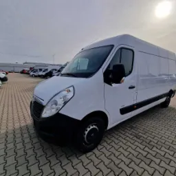Biały samochód dostawczy Opel Movano na brukowanym parkingu, widok z przodu i boku, w tle inne pojazdy i lekko pochmurne niebo.