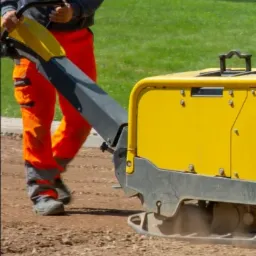 Operator w pomarańczowych spodniach roboczych obsługuje żółtą zagęszczarkę gruntu na placu budowy, przygotowując podłoże pod nawierzchnię.