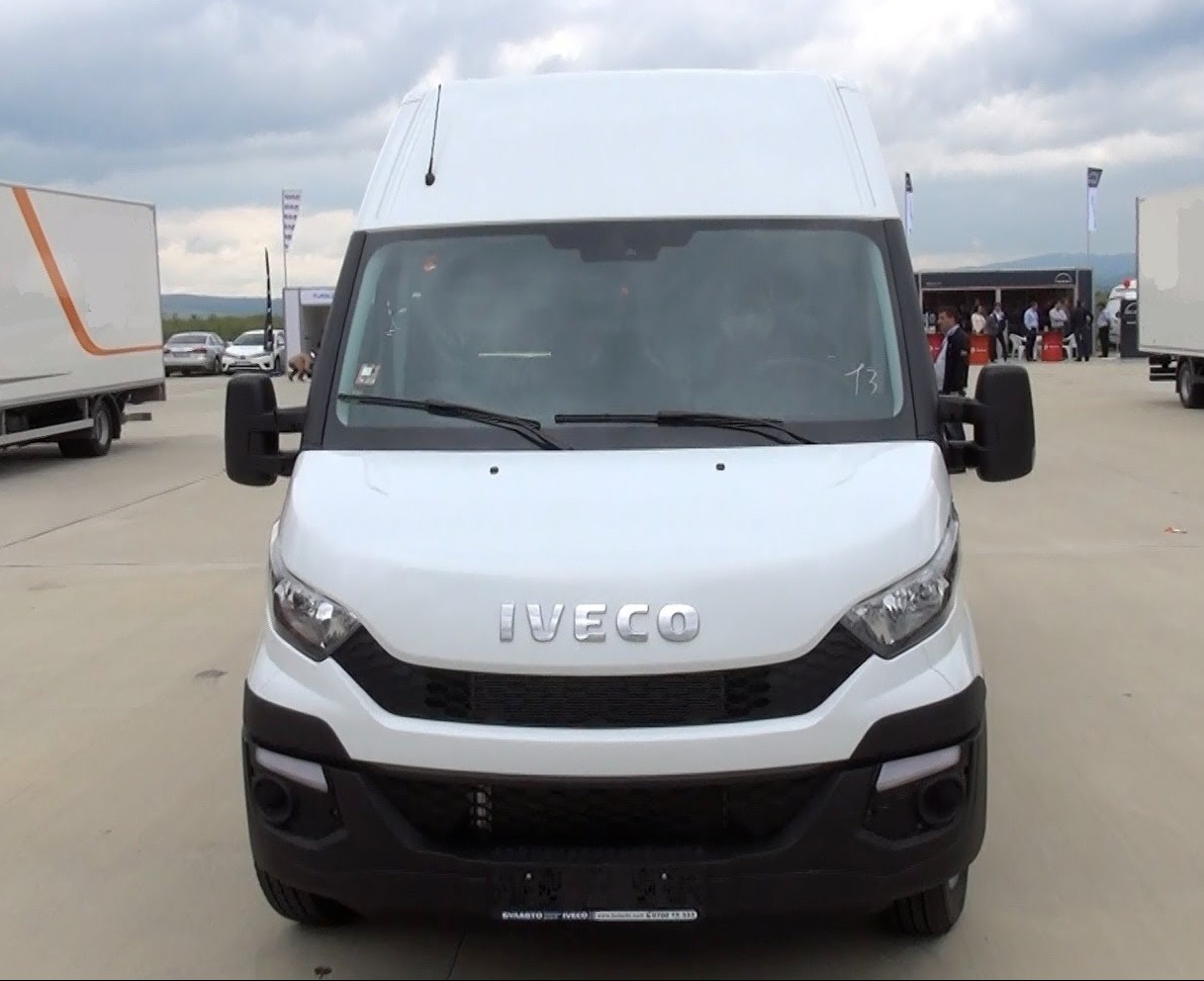 Frontowy widok białego samochodu dostawczego Iveco z anteną na dachu, zaparkowanego na betonowym placu podczas targów, z widocznymi innymi pojazdami i ludźmi w tle.