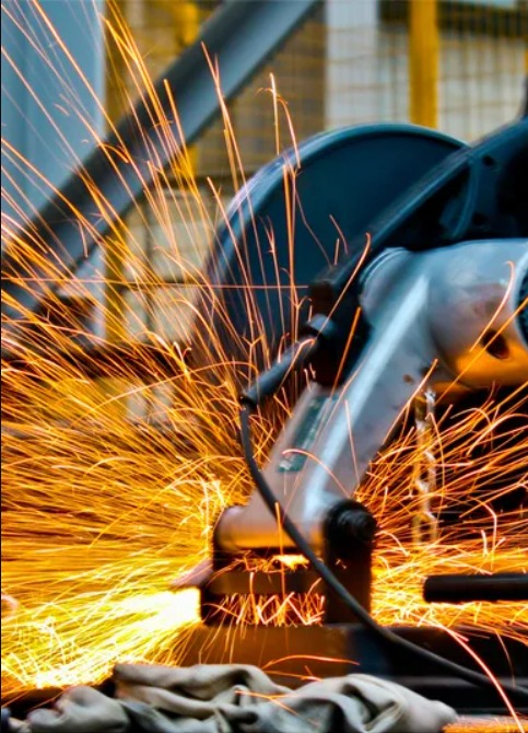 Metalowa piła tnąca podczas pracy, iskry rozbryzgują się w powietrzu, widoczne fragmenty konstrukcji stalowej w tle.