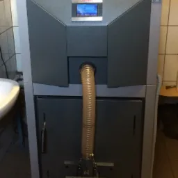 Szary piec na paliwo stałe marki DEFRO z widocznym panelem sterowania, manometrem i certyfikatem, zainstalowany w kotłowni z beżowymi płytkami.