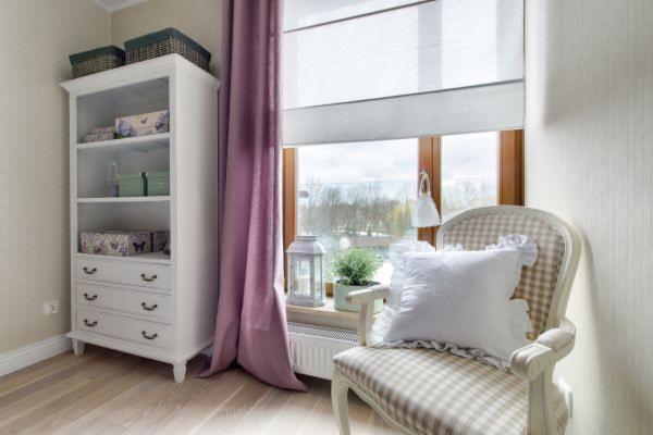 Jasny pokój z białym regałem wypełnionym dekoracyjnymi pudełkami, krzesłem w kratę z białą poduszką przy oknie z roletą rzymską i fioletową zasłoną.