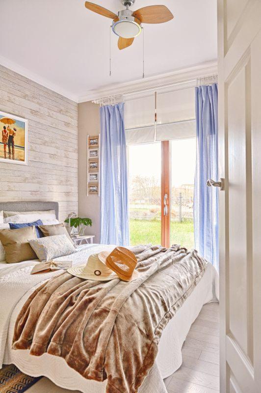 Jasna sypialnia z drewnianym panelem na ścianie, beżowym kocem na łóżku i słomkowymi kapeluszami, oświetlona sufitowym wentylatorem z drewnianymi łopatkami.
