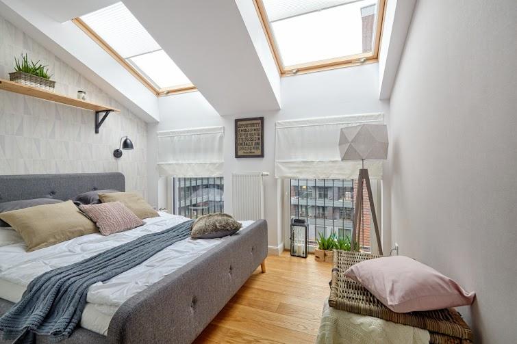 Sypialnia na poddaszu z szarym łóżkiem, drewnianą podłogą i oknami dachowymi z roletami, dekorowana roślinami w doniczkach i lampą podłogową z abażurem geometrycznym.