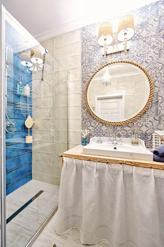 Jasna łazienka z niebieskimi akcentami: kabina prysznicowa z półkami na kosmetyki, umywalka z zasłonką z lnu, lustro w ramie z liny i wzorzyste płytki na ścianie.