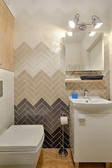 Nowoczesna łazienka z umywalką na szafce i toaletą, ściana wyłożona kafelkami w jodełkę w odcieniach beżu i brązu, oświetlenie punktowe.