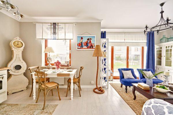 Jasne wnętrze jadalni i salonu w stylu marynistycznym, z białym stołem, drewnianymi krzesłami, zegarem stojącym, lampą z jutowego sznura, niebieskimi fotelami i zasłonami.