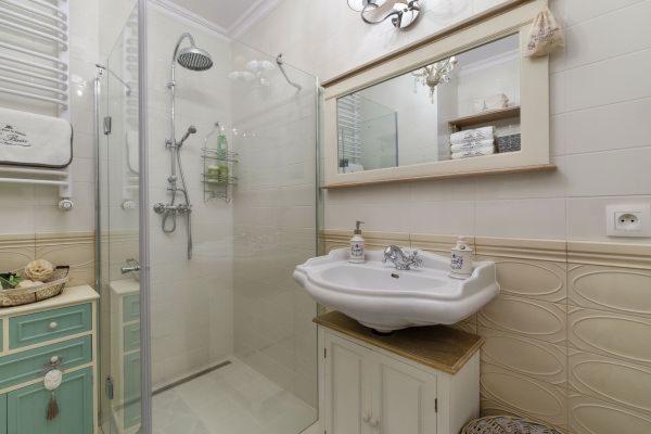Jasna łazienka z kabiną prysznicową, umywalką na szafce i dużym lustrem w ozdobnej ramie, widoczna dekoracyjna armatura i detale.
