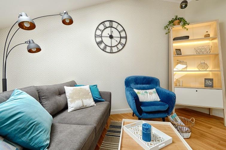 Salon z szarą sofą, niebieskim fotelem, poduszkami z motywem drzewa i geometrycznym wzorem, metalowym zegarem na ścianie oraz regałem z podświetleniem LED.