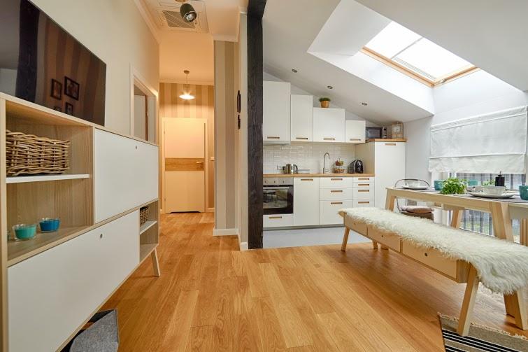 Jasne, przestronne wnętrze mieszkania na poddaszu z drewnianą podłogą, białymi meblami kuchennymi, stołem jadalnym z futrzaną ławką i widocznym przejściem do pokoju z telewizorem.