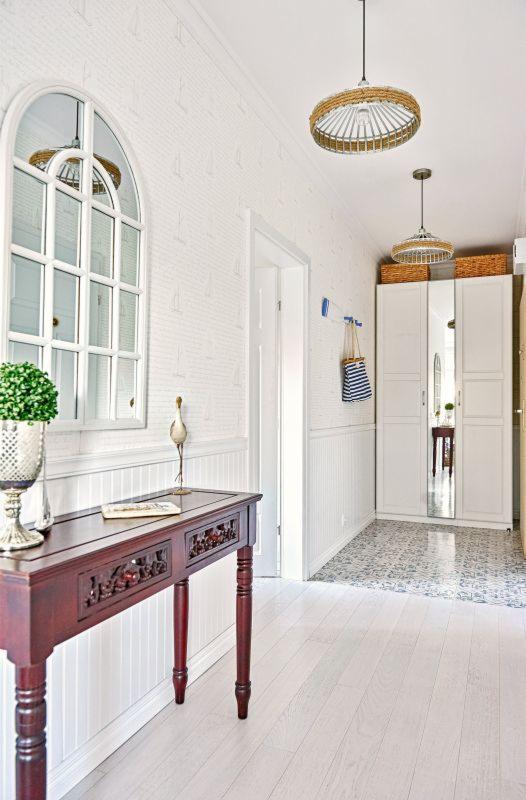 Jasny przedpokój w stylu marynistycznym z białymi ścianami w delikatny wzór żaglówek, drewnianą konsolą z rzeźbieniami, okrągłym lustrem w kształcie okna i lampami z elementami liny.