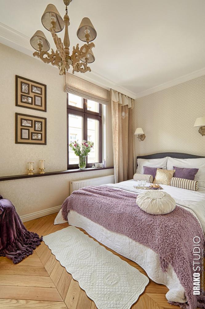 Przytulna sypialnia w stylu klasycznym z drewnianą podłogą, fioletową narzutą na łóżku, dekoracyjną lampą wiszącą, oknem z roletą rzymską i kwiatami oraz ramkami ze zdjęciami na ścianie.
