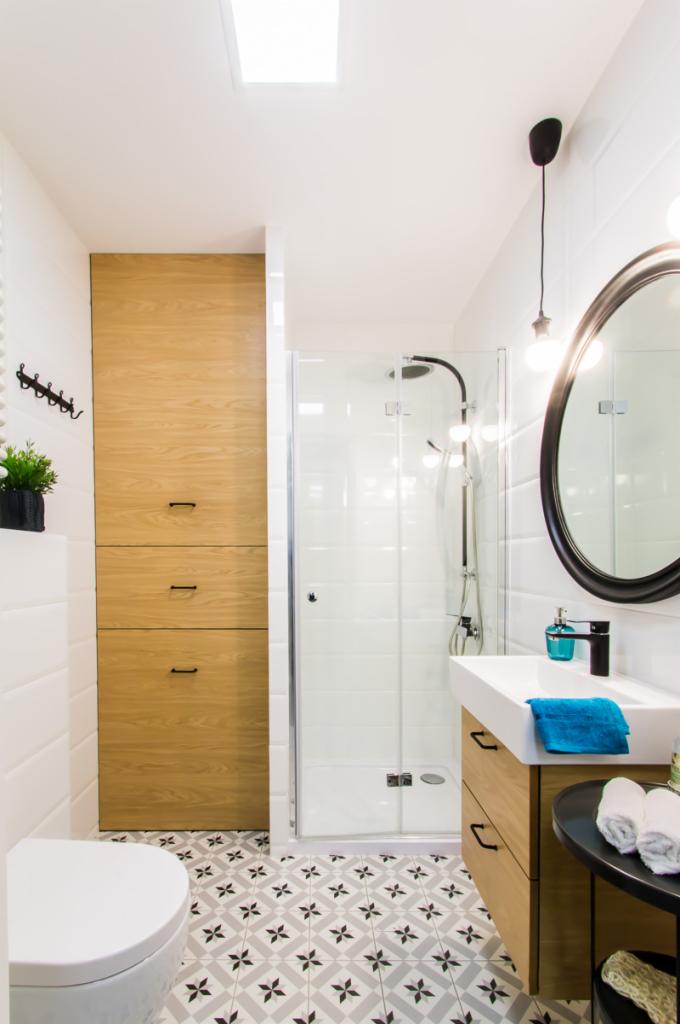 Nowoczesna łazienka z drewnianymi szafkami, kabiną prysznicową z czarną armaturą i wzorzystą podłogą w odcieniach szarości, oświetlona światłem dziennym z okna dachowego.