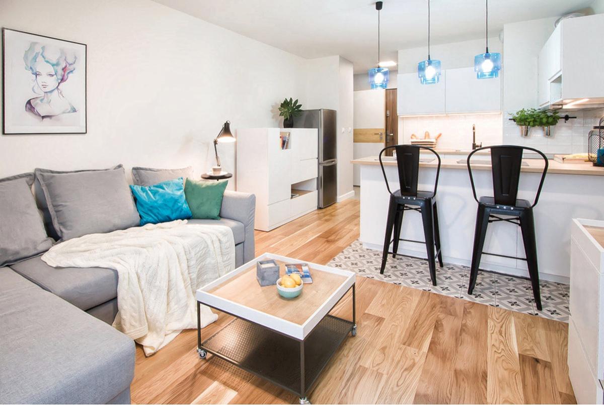 Jasne, nowoczesne wnętrze mieszkania z drewnianą podłogą, szarą sofą z turkusową poduszką, stolikiem kawowym na kółkach, białą zabudową kuchenną i niebieskimi lampami wiszącymi.