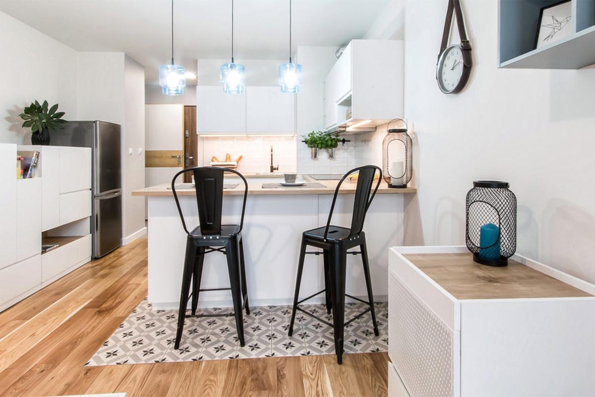 Nowoczesna, jasna kuchnia z drewnianą podłogą i wzorzystą posadzką pod wyspą, oświetlona trzema błękitnymi lampami wiszącymi, z dwoma czarnymi hokerami przy blacie.