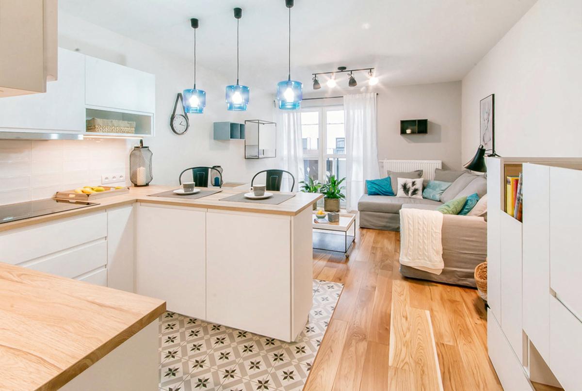 Jasne, przestronne wnętrze mieszkania z aneksem kuchennym, drewnianym blatem i niebieskimi lampami wiszącymi, widok na salon z szarą sofą i dekoracyjnymi poduszkami.