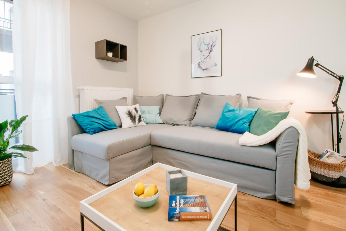 Jasnoszara sofa narożna z turkusowymi i szarymi poduszkami w salonie z drewnianą podłogą, stolikiem kawowym z cytrynami i książką o skandynawskim designie.
