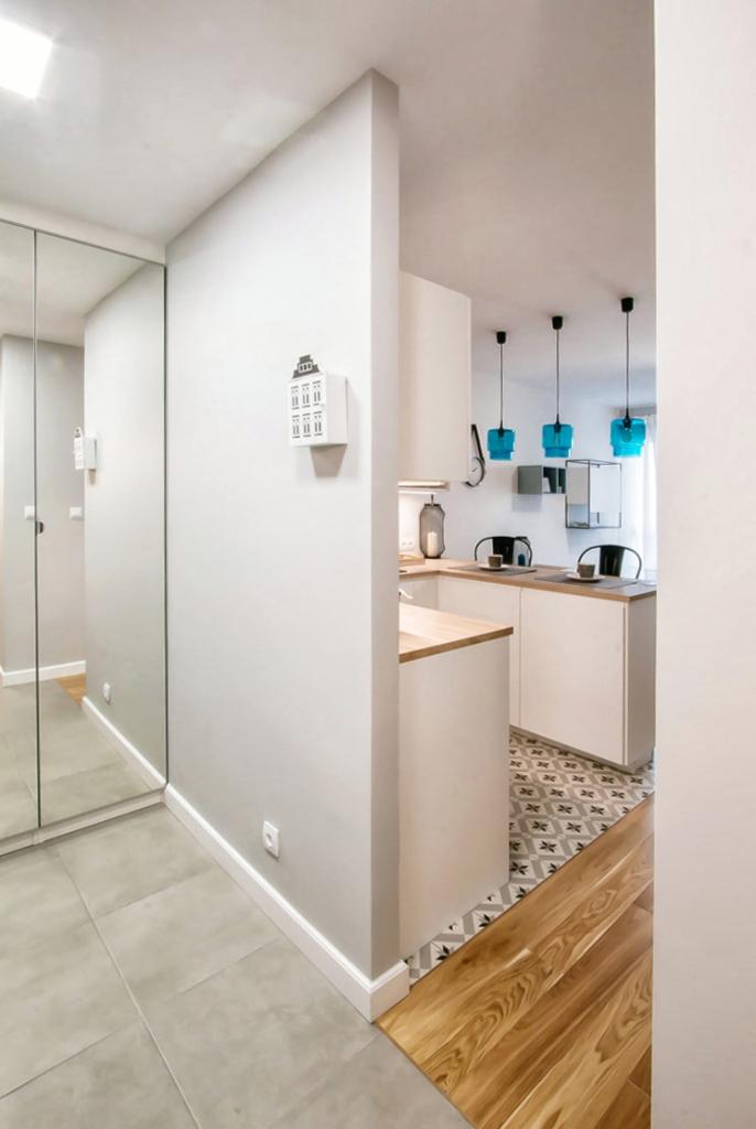 Nowoczesne wnętrze mieszkania: przedpokój z lustrzaną szafą i wejściem do kuchni z turkusowymi lampami wiszącymi, białą zabudową i wzorzystą podłogą.