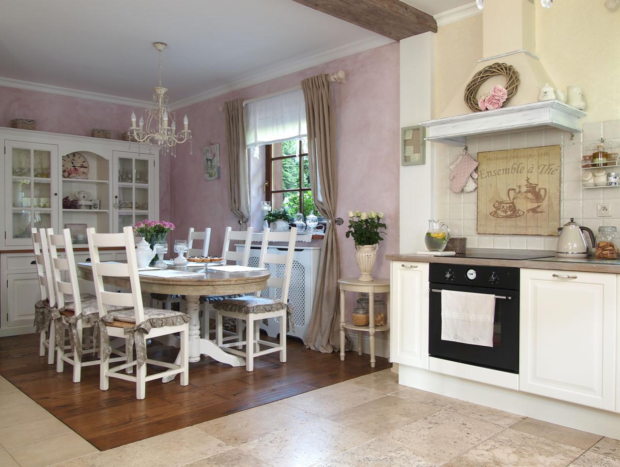 Jasna kuchnia i jadalnia w stylu rustykalnym z okrągłym drewnianym stołem, białymi krzesłami z poduszkami w kwiaty, kredensem z przeszklonymi drzwiczkami i dekoracyjnym okapem z wiankiem