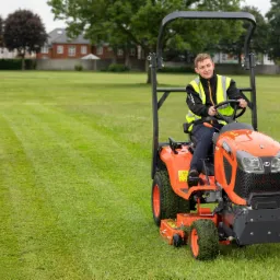 Operator w kamizelce odblaskowej na pomarańczowym traktorku do koszenia trawy Kubota R261, świeżo skoszony trawnik z widocznymi pasami, budynki w tle.