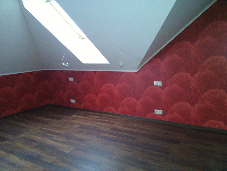 Poddasze z czerwonymi ścianami o fakturze kwiatowej i ciemnymi panelami podłogowymi, widoczne gniazdka elektryczne i okno dachowe.