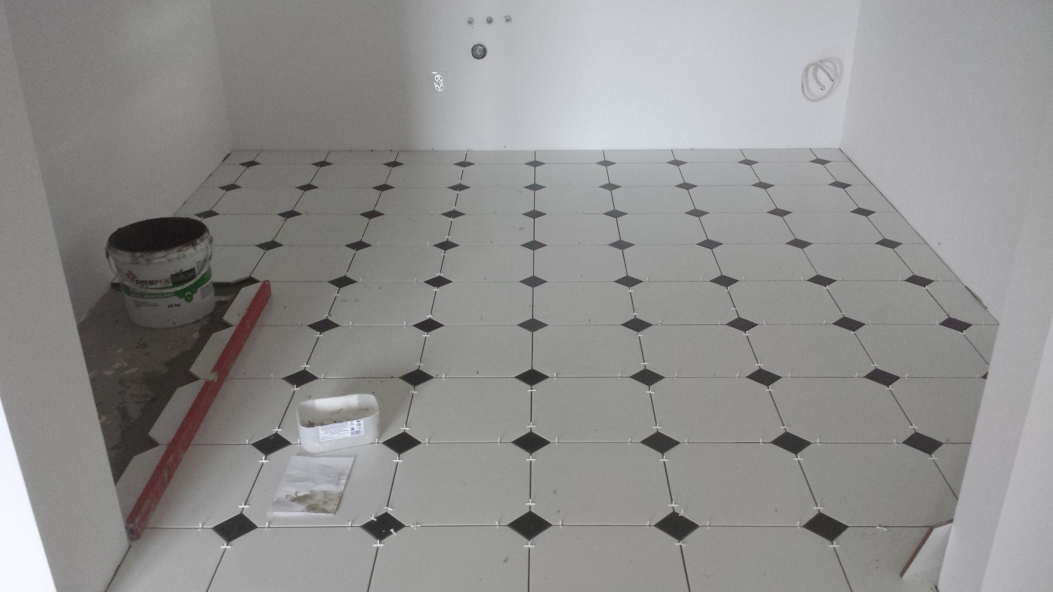 Układanie płytek podłogowych w łazience, widoczny kubeł z klejem, poziomica i pojemnik z krzyżykami dystansowymi.