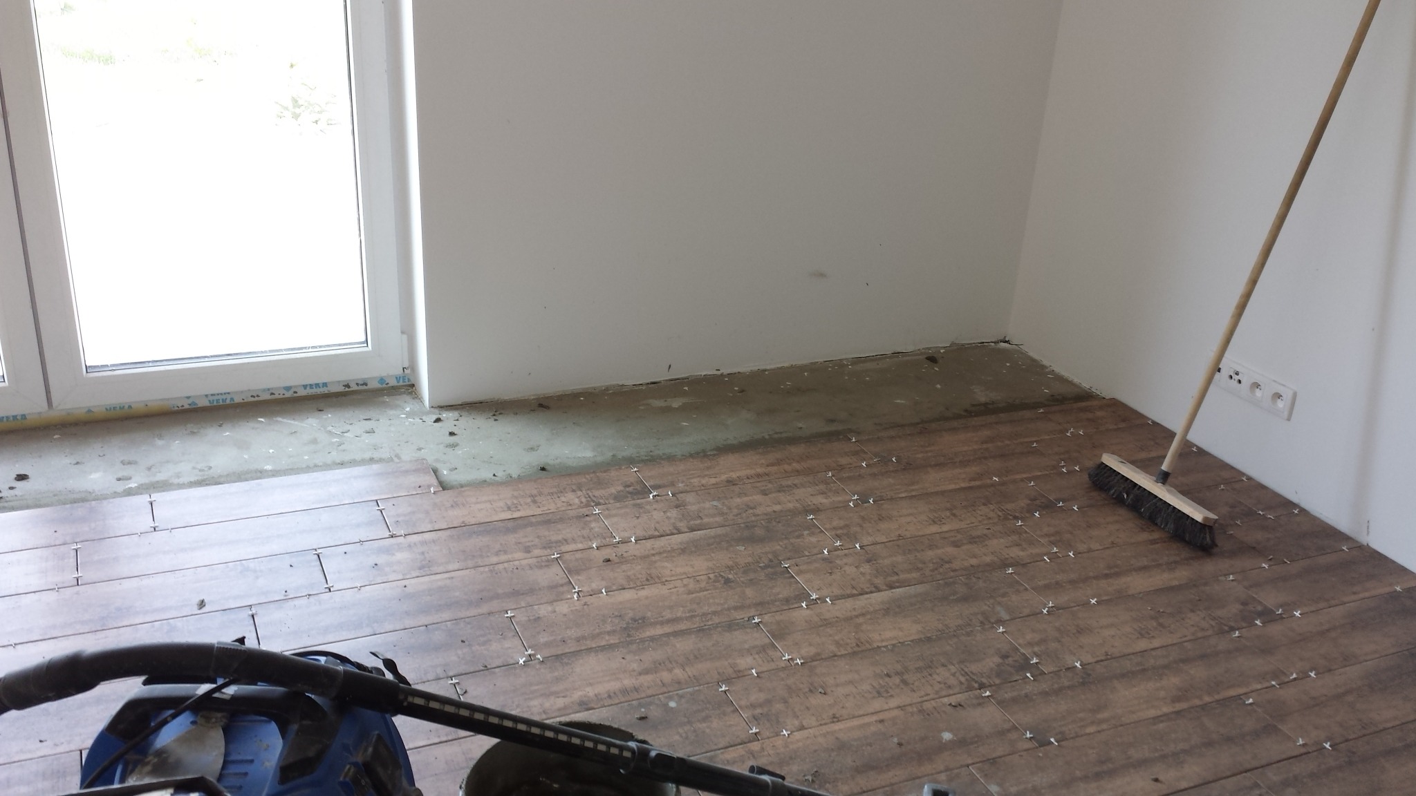 Układanie płytek imitujących drewno w pomieszczeniu, widoczne krzyżyki dystansowe, odkurzacz i szczotka, fragment podłogi bez płytek przy oknie.