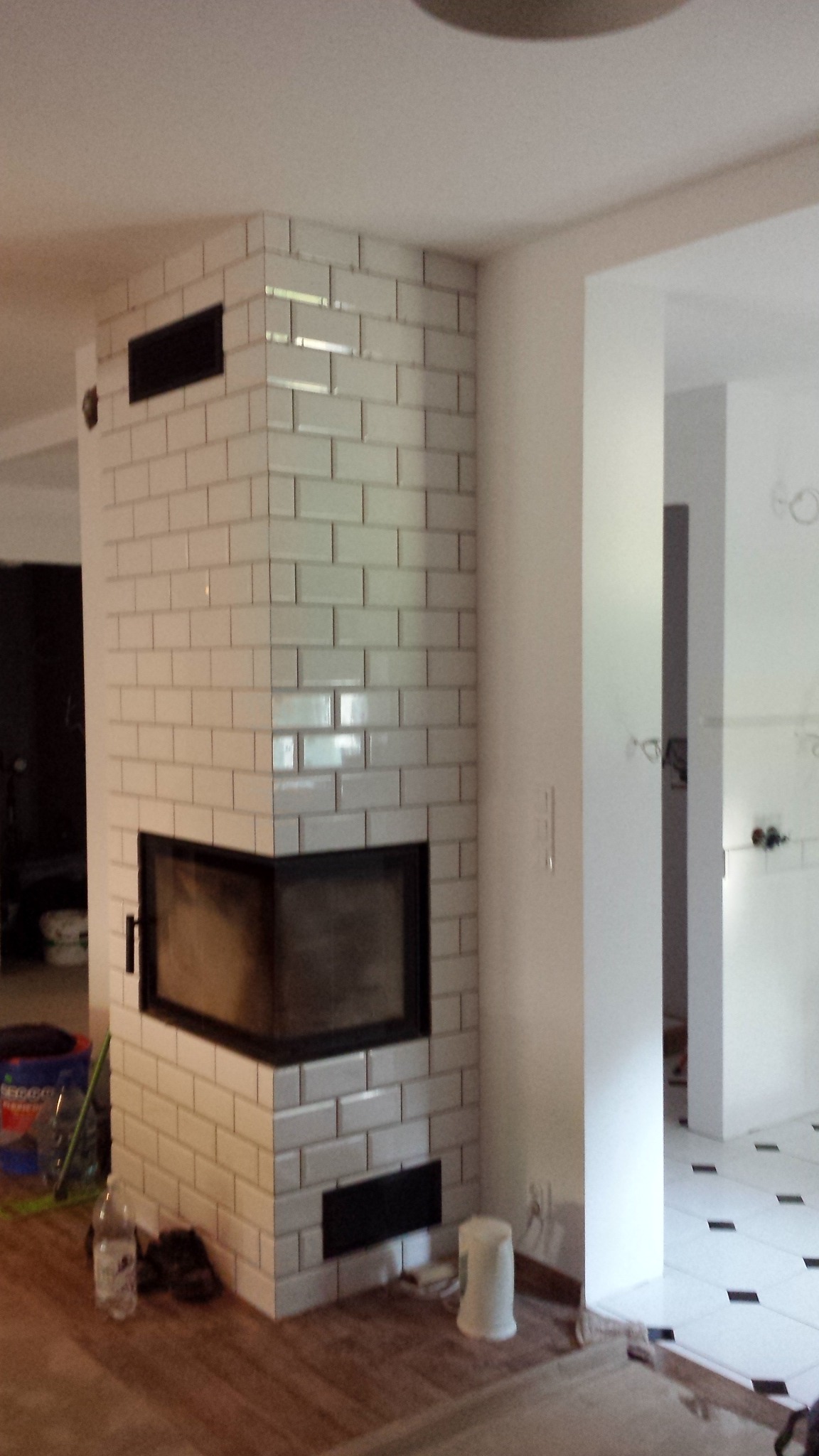 Obudowa kominka wykończona białymi, prostokątnymi płytkami ceramicznymi, z czarnym wkładem kominkowym i kratkami wentylacyjnymi, widok z boku, w tle białe ściany i podłoga z czarnymi akcentami.