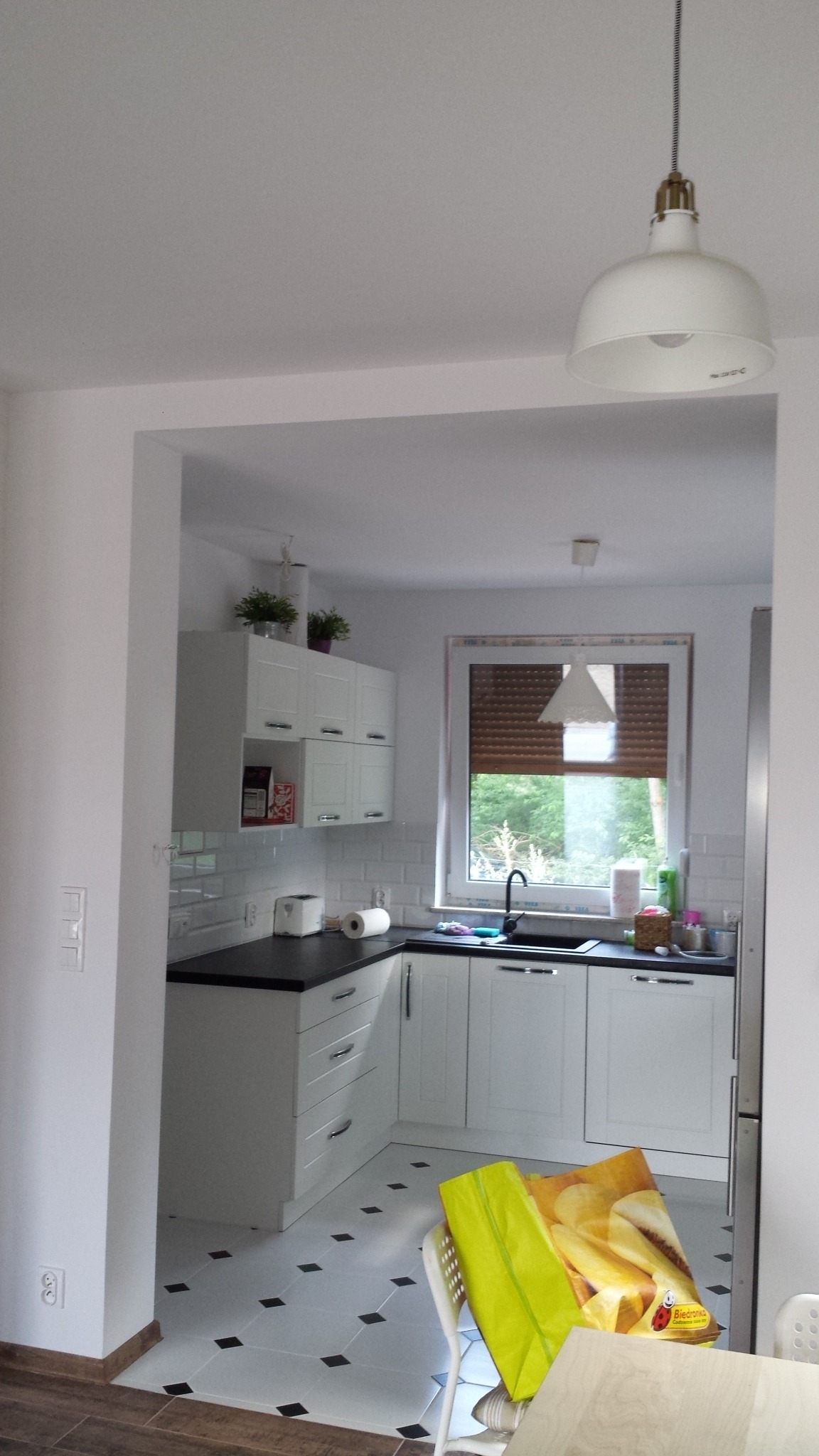 Jasna kuchnia z białymi szafkami i czarnym blatem w Pobiedziskach, z widocznym fragmentem podłogi wyłożonej białymi płytkami z czarnymi akcentami, oświetlona wiszącą lampą z białym kloszem.