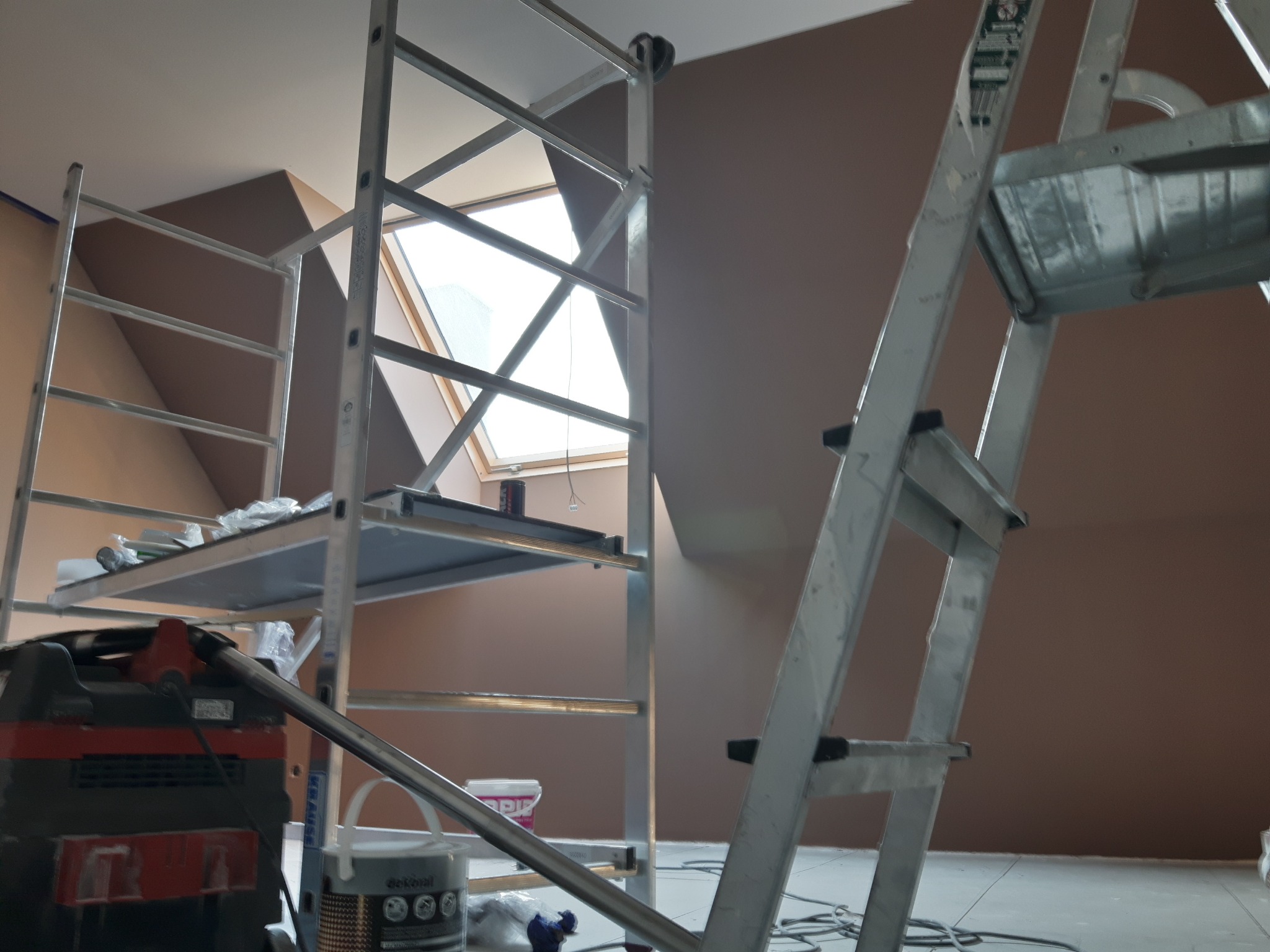 Pokój na poddaszu w trakcie malowania na brązowo, widoczne rusztowania, okno dachowe i odkurzacz przemysłowy.