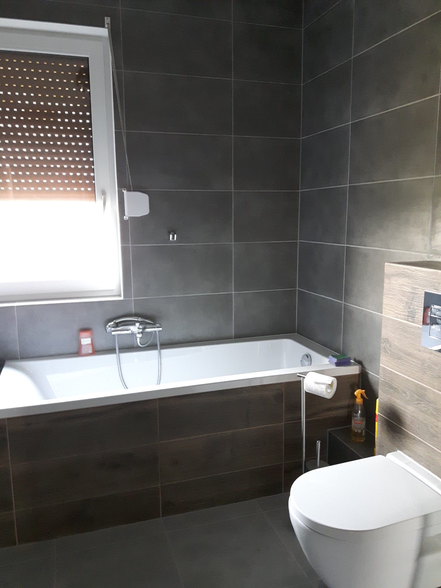 Nowoczesna łazienka z ciemnymi płytkami, białą wanną obudowaną drewnopodobną okładziną i wiszącą toaletą, okno z roletami w tle.