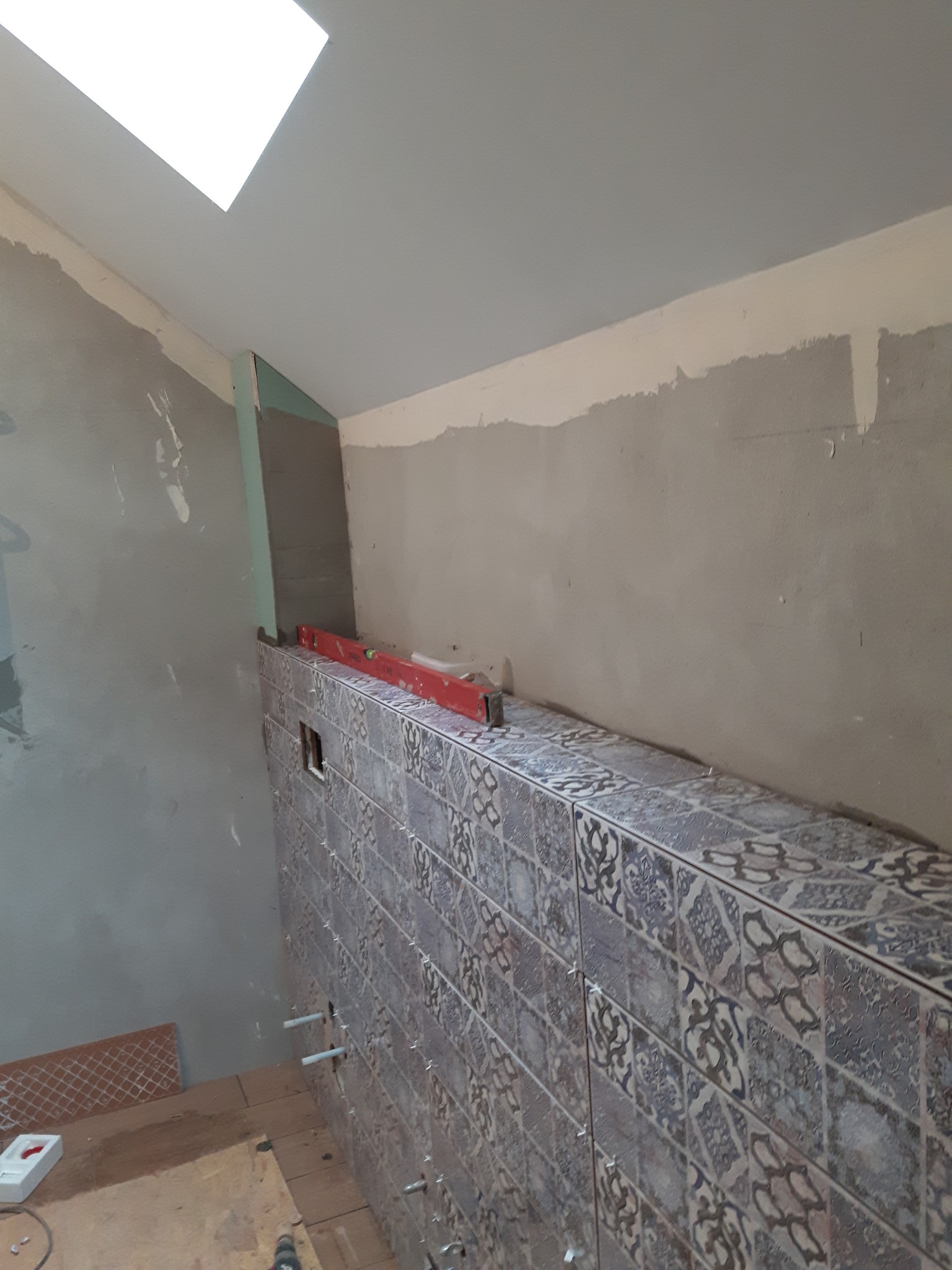 Montaż dekoracyjnych płytek ściennych w łazience z widoczną poziomicą, szpachlowaną ścianą i oknem dachowym.