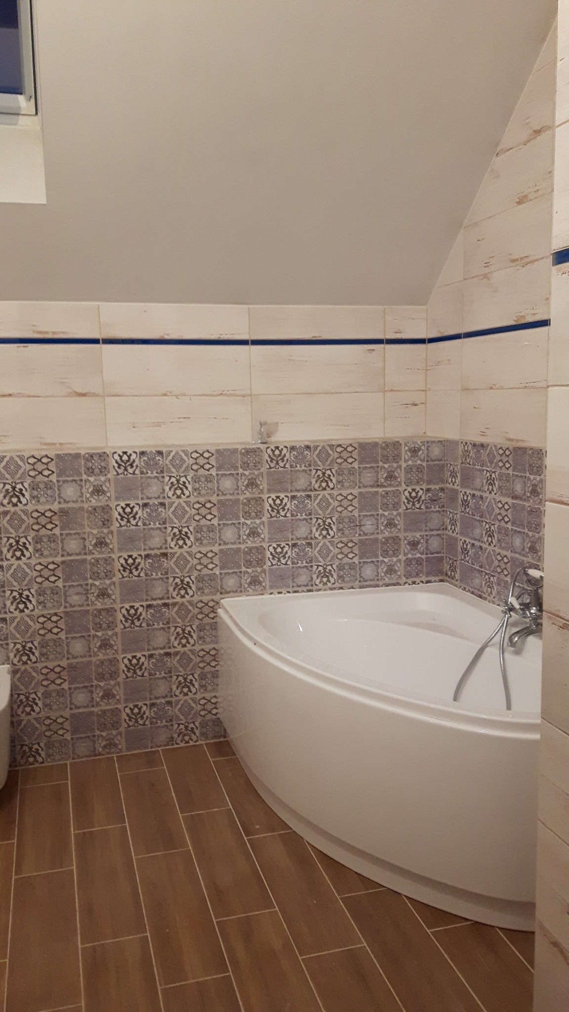 Wykończona łazienka na poddaszu z narożną wanną, płytkami w odcieniach beżu i brązu oraz dekoracyjnymi wzorami na ścianie, widoczne brązowe panele podłogowe imitujące drewno.